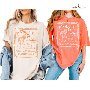 May include: Two t-shirts, one cream and one coral, with a graphic design. The design features the text "A Spritz Older" with a cocktail illustration, a cake, and the text "Olivia's 40th Birthday, Charleston, SC."