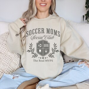 May include: A beige sweatshirt with the text "Soccer Moms Social Club" and a crest featuring a soccer ball, goal, chair, and coffee cup. The crest is surrounded by a laurel wreath and the text "The Real MVPs".