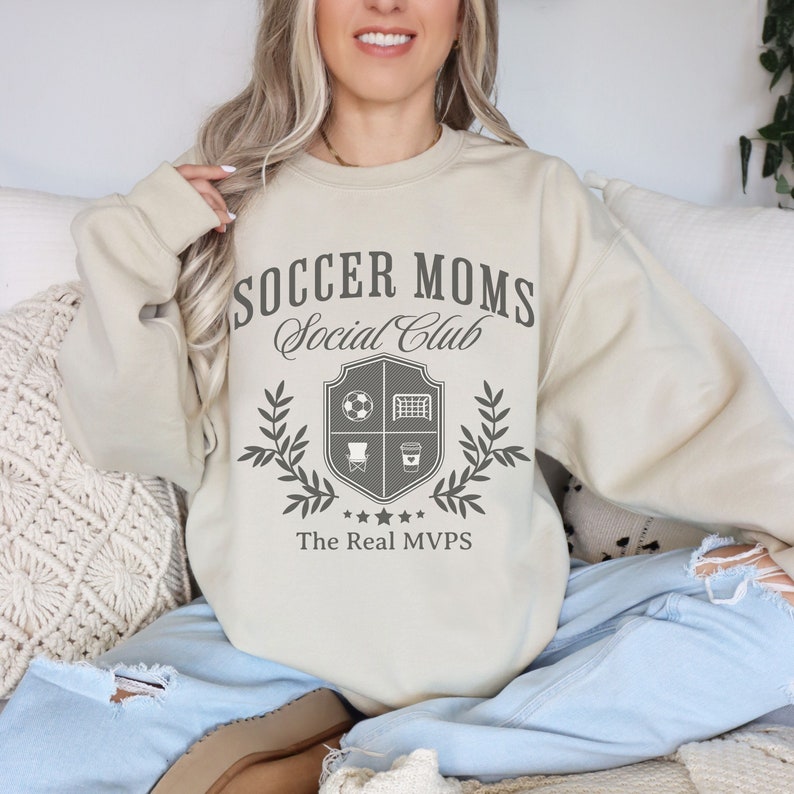 May include: A beige sweatshirt with the text "Soccer Moms Social Club" and a crest featuring a soccer ball, goal, chair, and coffee cup. The crest is surrounded by a laurel wreath and the text "The Real MVPs".
