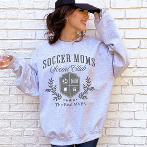 May include: Gray sweatshirt with the text "Soccer Moms Social Club" and a crest with a soccer ball, goal, chair, and coffee cup. The text "The Real MVPs" is below the crest.