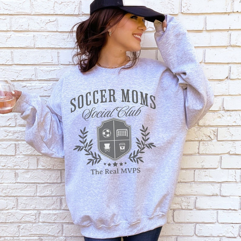May include: Gray sweatshirt with the text "Soccer Moms Social Club" and a crest with a soccer ball, goal, chair, and coffee cup. The text "The Real MVPs" is below the crest.