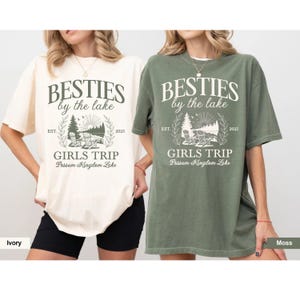 K&ouml;nnte beinhalten: Zwei T-Shirts, eines in Elfenbein und eines in Moosgr&uuml;n, mit dem Text "BESTIES by the lake" und "GIRLS TRIP Possum Kingdom Lake". Das Design zeigt eine Seeszene mit St&uuml;hlen, B&auml;umen und dem Jahr 2025. Die Shirts sind kurz&auml;rmelig.