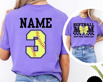 May include: Purple t-shirt with the text "NAME" and the number "3" in a softball design. The number is yellow with red stitching. The t-shirt is shown on a model. A second image shows a purple t-shirt with the text "SOFTBALL MOM" in black with a yellow softball and bow. The text "CRAZY PROUD. ALWAYS LOUD" is below the main text. The t-shirt is shown on a model. The text "VIOLET" is in a black circle.
