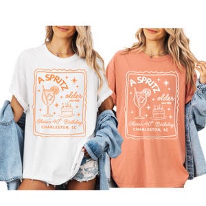 May include: Two t-shirts, one white and one coral, each featuring a rectangular design with the words "A Spritz" and "older estd 1985." The design includes a cocktail glass, a cake, and the text "Olivia's 40th Birthday, Charleston, SC."