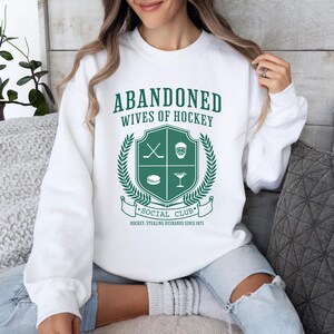 Puede incluir: Sudadera blanca con texto verde que dice "Abandoned Wives of Hockey" y un escudo verde con palos de hockey, una m&aacute;scara de hockey, un disco y una copa de martini. El texto debajo del escudo dice "Social Club" y "Hockey: Stealing Husbands Since 1875."