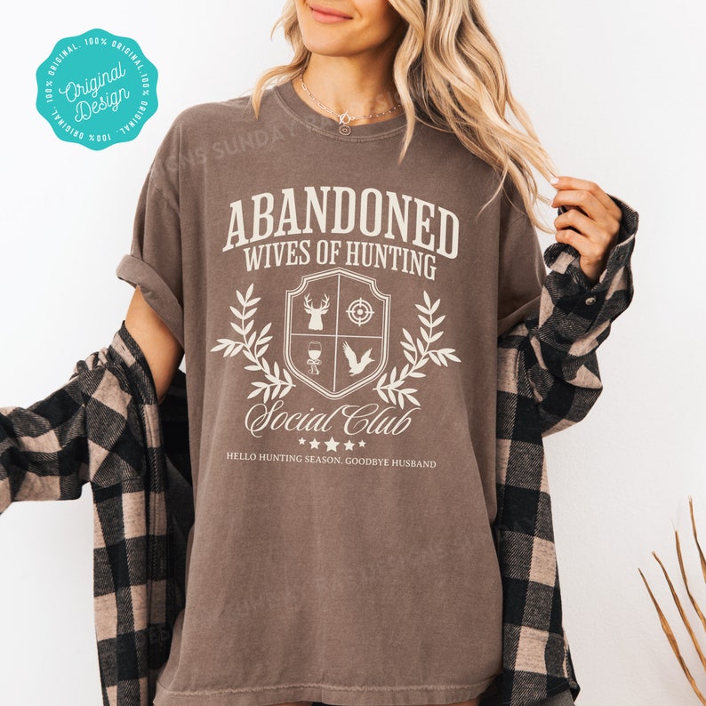 May include: A brown t-shirt with a white graphic design that says "Abandoned Wives of Hunting Social Club" with a hunting-themed crest and the text "Hello Hunting Season, Goodbye Husband".