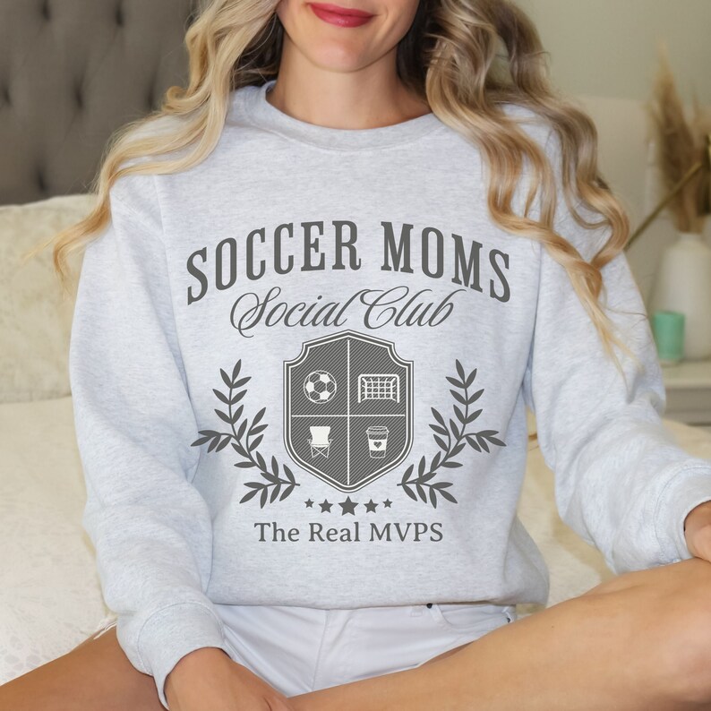 May include: Gray sweatshirt with the text "Soccer Moms Social Club" and a crest featuring a soccer ball, goal, chair, and coffee cup. The text "The Real MVPs" is below the crest.