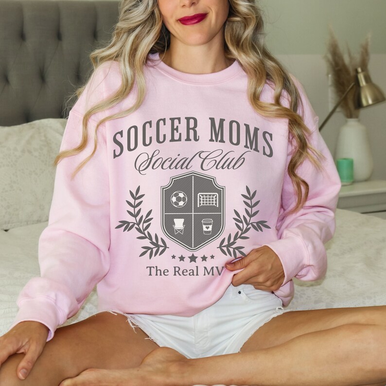May include: Pink sweatshirt with the text "Soccer Moms Social Club" and a crest featuring a soccer ball, goal, chair, and coffee cup. The text "The Real MV" is below the crest.