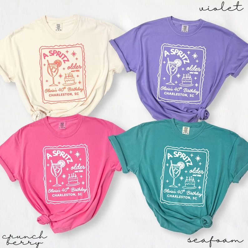 May include: Four t-shirts in cream, violet, pink, and teal. Each shirt features a white graphic with the text "A Spritz + older" and "Olivia's 40th Birthday, Charleston, SC" with a cocktail and cake illustration.