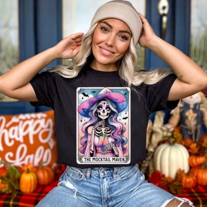May include: A black t-shirt with a colorful illustration of a skeleton witch holding a cocktail. The illustration is framed in a card with the text "The Mocktail Maven".