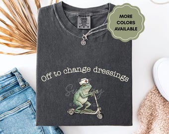 Puede incluir: Camiseta gris oscuro con la frase blanca "Off to change dressings". La camiseta presenta una rana de dibujos animados con gorro de enfermera montada en un patinete. También se ve un collar plateado con un colgante de sol.