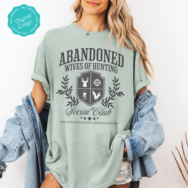 May include: A light green t-shirt with the text "Abandoned Wives of Hunting Social Club" and a crest featuring a deer, a target, a wine glass, and a bird. The text "Hello Hunting Season. Goodbye Husband" is printed below the crest.