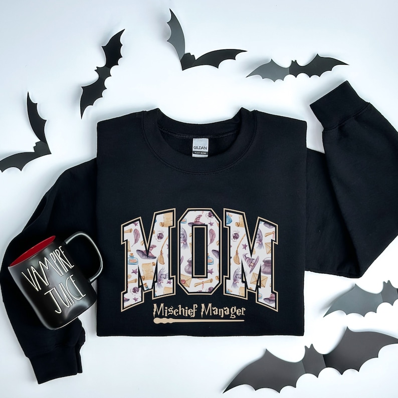 May include: Black sweatshirt with a gold "MOM" graphic featuring a pattern of witch hats, brooms, and other Halloween-themed items. The text "Mischief Manager" is below the graphic.