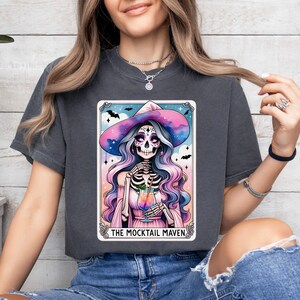 May include: A dark gray t-shirt with a colorful illustration of a skeleton witch holding a cocktail. The illustration is framed in a card-like border with the text "The Mocktail Maven".