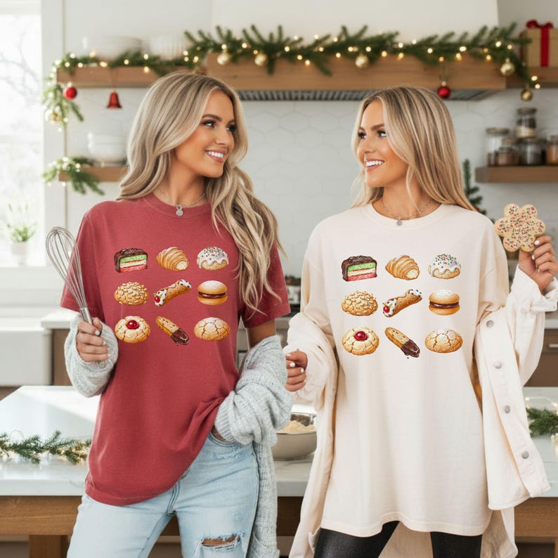 May include: Two women wearing t-shirts with a watercolor illustration of various pastries. One shirt is maroon, the other is cream-colored. The pastries include cookies, croissants, and cannoli. One woman holds a whisk, the other a cookie.
