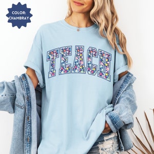 May include: A light blue t-shirt with the word "TEACH" in large, navy blue letters. The letters are filled with a colorful floral pattern.