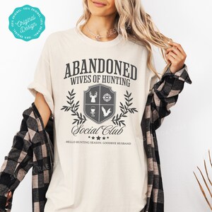 May include: A beige t-shirt with a black graphic that says "Abandoned Wives of Hunting Social Club" with a hunting-themed crest and the text "Hello Hunting Season, Goodbye Husband".