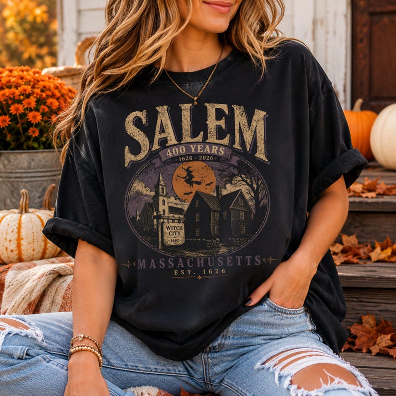 May include: A black short-sleeve t-shirt featuring a graphic print with the text SALEM 400 YEARS 1626-2026 MASSACHUSETTS EST. 1626. The design includes a silhouette of a witch on a broomstick, a church, houses, and a sign reading WITCH CITY 1692.