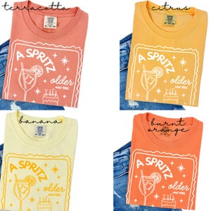 May include: Four t-shirts in terracotta, citrus, banana, and burnt orange. Each shirt features a white graphic with a cocktail, text that reads "A Spritz Older estd 1985", and a cake. The shirts are displayed on denim.