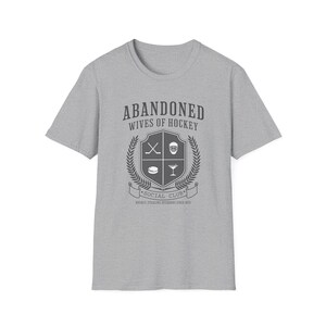 Pu&ograve; includere: Una maglietta grigia con un grafico di uno stemma che dice "Abandoned Wives of Hockey" e "Social Club" con icone di bastone da hockey, disco e bicchiere di martini.