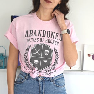 Pu&ograve; includere: Maglietta rosa con il testo "ABANDONED WIVES OF HOCKEY" e uno stemma che presenta bastoni da hockey, una maschera da hockey, un disco e un bicchiere da martini. Lo stemma &egrave; circondato da una corona di alloro e dal testo "SOCIAL CLUB".