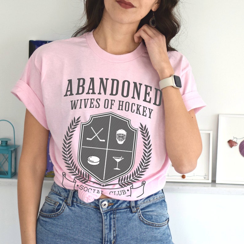 Pu&ograve; includere: Maglietta rosa con il testo "ABANDONED WIVES OF HOCKEY" e uno stemma che presenta bastoni da hockey, una maschera da hockey, un disco e un bicchiere da martini. Lo stemma &egrave; circondato da una corona di alloro e dal testo "SOCIAL CLUB".