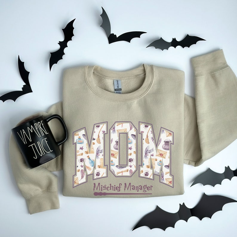 May include: A tan sweatshirt with a purple "MOM" graphic featuring a pattern of witchy items like brooms, potions, and hats. The text "Mischief Manager" is below the graphic with a purple wand.