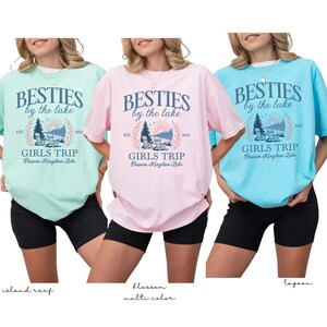 K&ouml;nnte beinhalten: Drei T-Shirts in den Farben Island Reef, Blossom und Lagoon. Jedes Shirt tr&auml;gt den Text "BESTIES by the lake" &uuml;ber "GIRLS TRIP" und "Possom Kingdom Lake". Die Shirts sind kurz&auml;rmelig und werden mit schwarzen Shorts getragen.
