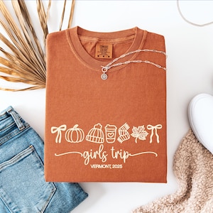 May include: Rust-colored Comfort Colors t-shirt with a white graphic of fall icons and the words "girls trip Vermont, 2025." A silver necklace and a pair of blue jeans are next to the shirt.
