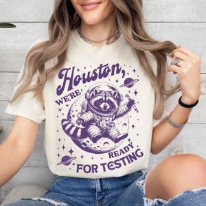 May include: A white t-shirt with a purple graphic of a raccoon wearing an astronaut suit and floating on a moon. The text "Houston, we're ready for testing" is printed on the shirt.