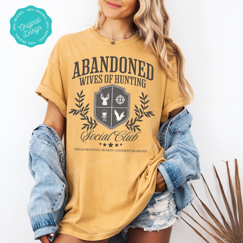 May include: A mustard yellow t-shirt with the text "Abandoned Wives of Hunting Social Club" in black. The shirt has a gray shield with a deer, target, wine glass, and bird inside. The text "Hello Hunting Season. Goodbye Husband" is below the shield.