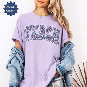 May include: A lavender t-shirt with the word "TEACH" printed in a floral pattern. The letters are outlined in navy blue.