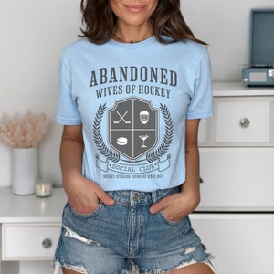 Pu&ograve; includere: Maglietta blu chiaro con una grafica grigia di uno stemma. Lo stemma ha una mazza da hockey, una maschera da hockey, un disco e un bicchiere da martini. Il testo sulla maglietta recita "Abandoned Wives of Hockey Social Club Hockey Stealing Husbands Since 1975."