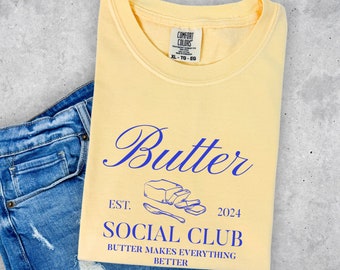 May include: A pale yellow t-shirt with the word "Butter" in blue script. Below, it reads "EST. 2024 SOCIAL CLUB BUTTER MAKES EVERYTHING BETTER" with a butter and knife graphic. The shirt is folded next to blue denim jeans.