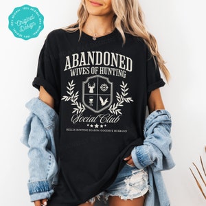 May include: Black t-shirt with white text that reads "Abandoned Wives of Hunting Social Club" with a crest featuring a deer, target, wine glass, and bird. The text below the crest reads "Hello Hunting Season, Goodbye Husband."