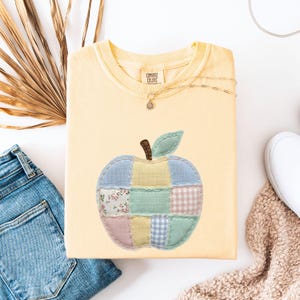 May include: A light yellow sweatshirt with a patchwork apple design. The apple is made of various pastel fabrics, including floral, gingham, and solid colors. The sweatshirt has a crew neck and long sleeves. The brand tag reads "Comfort Colors".