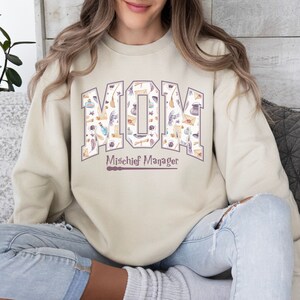 May include: A beige sweatshirt with a graphic print of the word "MOM" in purple letters. The letters are filled with a pattern of watercolor illustrations of magical objects, such as wands, potions, and feathers. The text "Mischief Manager" is printed below the word "MOM" in purple letters.