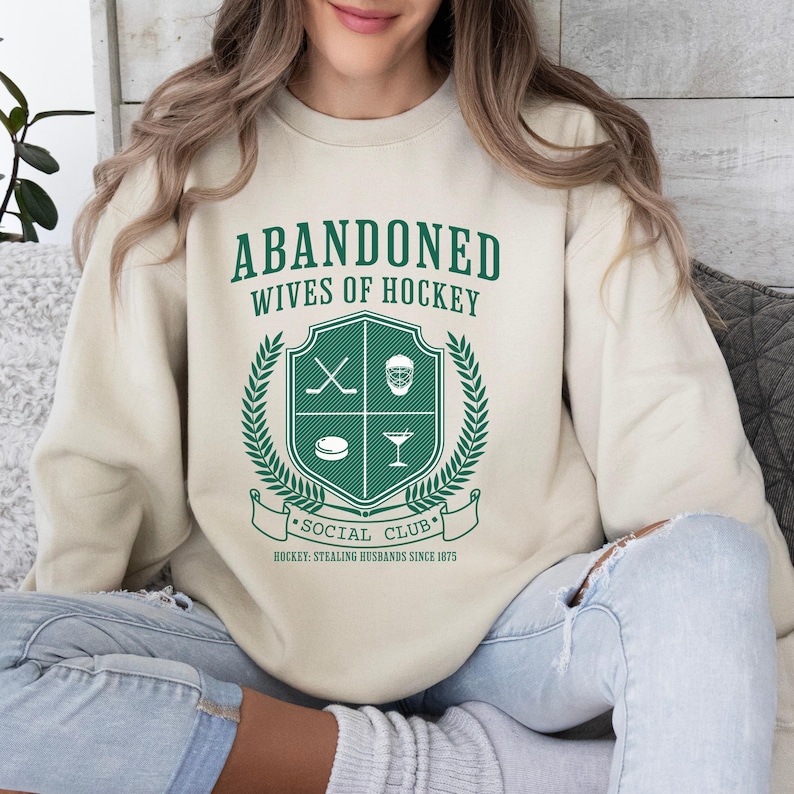 Puede incluir: Una sudadera beige con el texto "Abandoned Wives of Hockey" y un escudo verde con palos de hockey, una m&aacute;scara de hockey, un disco y una copa de martini. El escudo est&aacute; rodeado de una corona de laurel y una pancarta que dice "Social Club" y "Hockey. Stealing Husbands Since 1975."