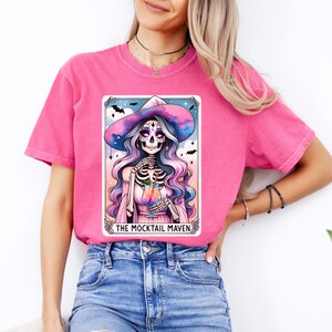 May include: A pink t-shirt with a graphic of a skeleton witch wearing a witch hat and holding a cocktail. The graphic is in a tarot card style with the text "THE MOCKTAIL MAVEN".