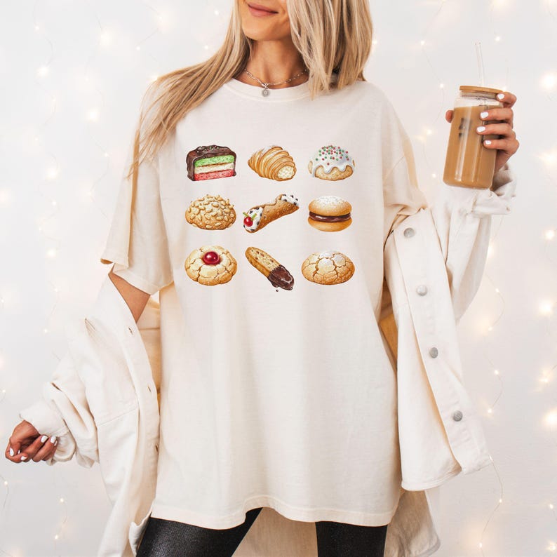 May include: Cream-colored t-shirt featuring a watercolor illustration of nine different pastries. The pastries include a croissant, a cannoli, and various cookies. The shirt is worn by a person holding a beverage in a clear glass with a wooden lid.