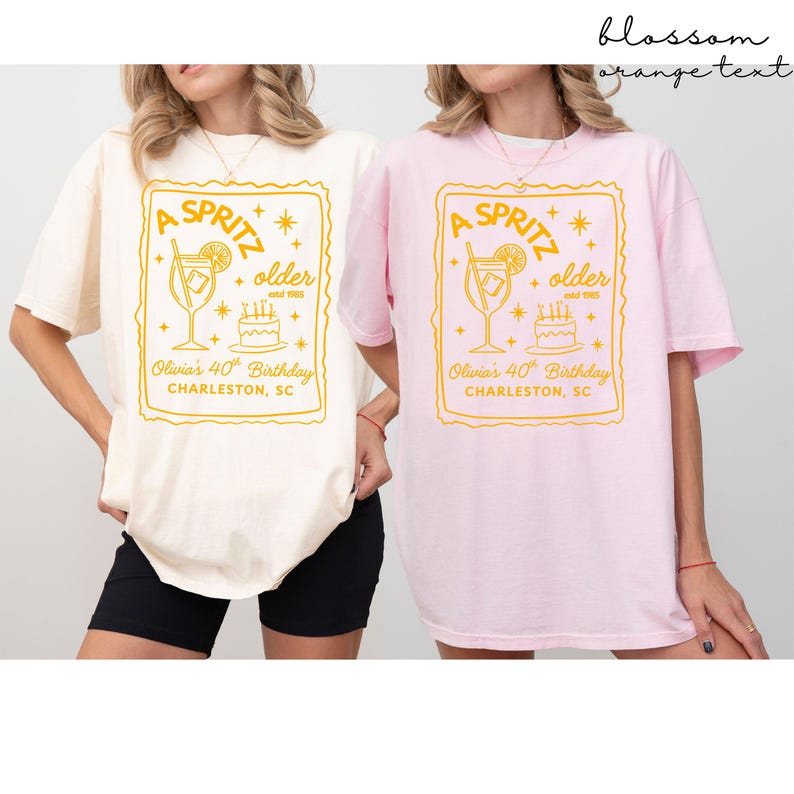 May include: Two oversized t-shirts, one cream and one pink, each featuring a yellow graphic. The graphic includes the text "A Spritz", "older", and "Olivia's 40th Birthday, Charleston, SC", along with a cocktail illustration and a cake.