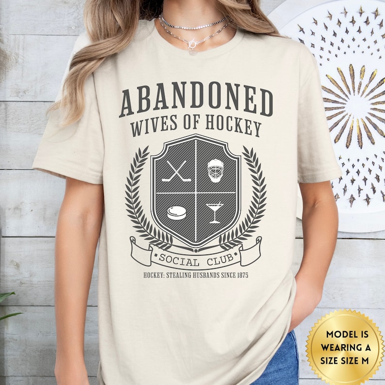 Pu&ograve; includere: Una maglietta beige con uno stemma grigio che presenta bastoni da hockey, una maschera da hockey, un disco e un bicchiere da martini. Il testo "Abandoned Wives of Hockey - Social Club - Hockey: Stealing Husbands Since 1875" &egrave; stampato sulla maglietta.