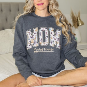 May include: A dark gray sweatshirt with the word "MOM" printed in large, white letters with a pattern of witchy items inside the letters. The text "Mischief Manager" is printed below the word "MOM" in a smaller font.