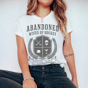 Pu&ograve; includere: Maglietta bianca con il testo "ABANDONED WIVES OF HOCKEY" e uno stemma con bastoni da hockey, una maschera da hockey, un disco e un bicchiere da martini. Il testo "SOCIAL CLUB" &egrave; sotto lo stemma con il testo "HOCKEY: STEALING HUSBANDS SINCE..." sotto.