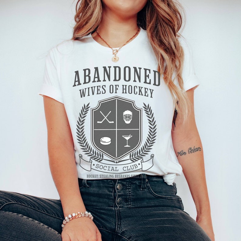 Pu&ograve; includere: Maglietta bianca con il testo "ABANDONED WIVES OF HOCKEY" e uno stemma con bastoni da hockey, una maschera da hockey, un disco e un bicchiere da martini. Il testo "SOCIAL CLUB" &egrave; sotto lo stemma con il testo "HOCKEY: STEALING HUSBANDS SINCE..." sotto.