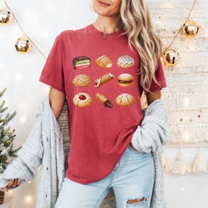 May include: A red t-shirt featuring a watercolor illustration of various pastries, including a croissant, a cannoli, and a cookie with a cherry. The shirt is a comfortable fit and has a crew neck.