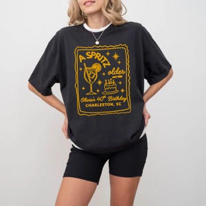 May include: Black t-shirt with a white collar featuring a yellow graphic. The graphic reads "A Spritz older and 1985" with a cocktail, cake, and text "Olivia's 40th Birthday Charleston, SC."