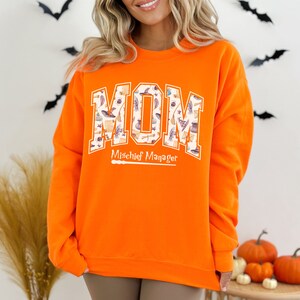 May include: Orange sweatshirt with a white graphic that says "MOM" with a pattern of witches hats, brooms, and stars inside the letters. The text "Mischief Manager" is below the graphic with a small wand underneath.