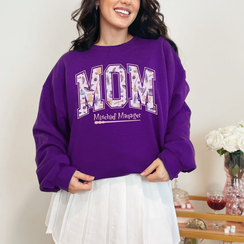 May include: Purple sweatshirt with the word "MOM" in white letters with a pattern of magical objects inside the letters. The text "Mischief Manager" is below the word "MOM" with a wand underneath.