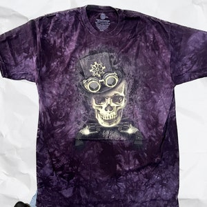 May include: Purple tie-dye t-shirt featuring a graphic of a skull wearing a top hat and goggles. The skull is in front of a steampunk-style machine.